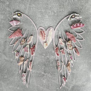 May include: Pink paper angel wings with a heart in the center. The wings are decorated with small pieces of pink and white china with floral patterns.