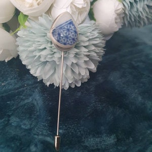 May include: A silver pin with a blue and white ceramic egg-shaped charm. The charm has a floral pattern.