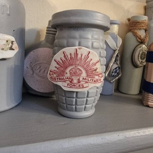 Puede incluir: Una botella de cerámica gris con una superficie texturizada y una etiqueta blanca que dice "Australian Military Forces".