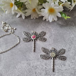 Könnte beinhalten: Zwei silberne Libellen-Anhänger mit floralen Akzenten. Die Libellen-Anhänger haben detaillierte Flügel und ein florales Design in der Mitte ihres Körpers. Eine Libelle hat eine rosa Blume und die andere eine gelbe Blume.