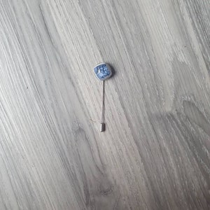 May include: A blue and white ceramic pin with a silver stem. The pin is shaped like a small, rounded rectangle.