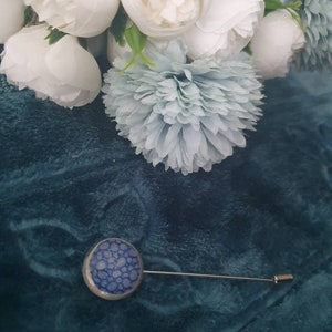 May include: A blue and white patterned pin with a silver pin back. The pin is on a blue fabric background.