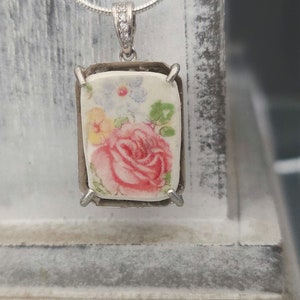 Peut inclure: Un collier en argent avec un pendentif rectangulaire orné d'un motif floral vintage. Le pendentif est en céramique blanche avec une rose rose et d'autres fleurs dans des tons de jaune, bleu et vert.
