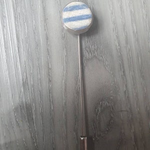 May include: A round lapel pin with a silver-colored pin and a circular head. The head features a white background with two horizontal light blue stripes. The pin is a decorative accessory.