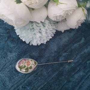 May include: A white porcelain pin with a floral design. The pin is oval shaped and has a silver pin back. The pin is on a blue background.