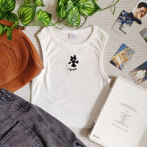 May include: White ribbed tank top with a black graphic of a creature and the letters "r p w p" below it. The top is sleeveless and has a round neckline. The image also includes a brown purse, gray jeans, and a book.