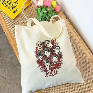 May include: Cream-colored tote bag with a graphic of seven people in sunglasses and red shirts, with the number "2.0" below. The bag has two handles and is filled with tulips. A yellow book with the days of the week is visible.
