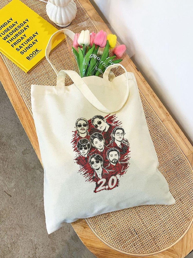 May include: Cream-colored tote bag with a graphic of seven people in sunglasses and red shirts, with the number "2.0" below. The bag has two handles and is filled with tulips. A yellow book with the days of the week is visible.