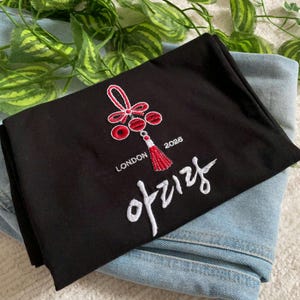 May include: Black folded t-shirt with a red and white embroidered design. The design includes a stylized knot, a tassel, and the text "LONDON 2026" and Korean characters. The t-shirt is resting on a pair of blue jeans.