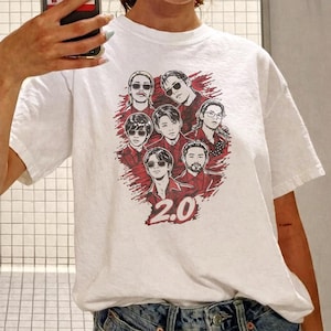 May include: White t-shirt featuring a graphic of seven people in red and black. The graphic includes the number "2.0" at the bottom. The shirt is made of a soft material.