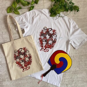 May include: A white t-shirt and a beige tote bag with a graphic of a group of people in sunglasses and the text "FIRST STEP IN ELIER 2.0". A colorful fan with a black handle is also visible.