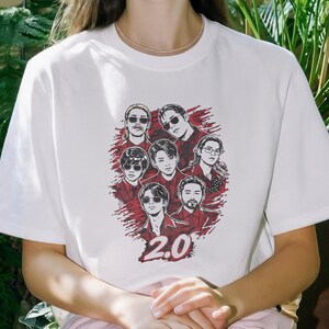 May include: White t-shirt featuring a graphic of seven people with sunglasses and the number "2.0" in red. The design is set against a red splatter background. The shirt is a casual wear item.