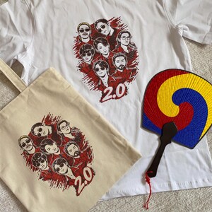 May include: White t-shirt and canvas tote bag featuring a graphic of a group of people with sunglasses and the number "2.0" in red. A traditional Korean fan with red, yellow, and blue colors is also visible.