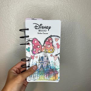 May include: A Disney-themed savings challenge notebook with a spiral binding. The cover features a Minnie Mouse bow, a castle illustration, and the Disney logo. The notebook is designed to help save $5000.