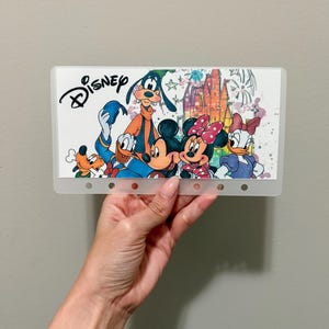 Op de afbeelding: Een doorzichtige plastic envelop met een kleurrijk Disney-ontwerp. Het ontwerp bevat Mickey Mouse, Minnie Mouse, Goofy, Donald Duck en Pluto, met het Disney-logo bovenaan. De achtergrond toont een kasteel en vuurwerk.