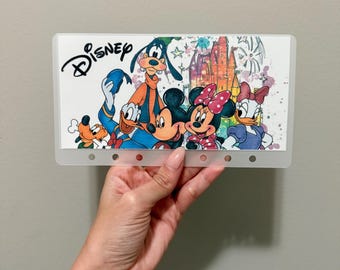 Könnte beinhalten: Eine transparente Kunststoffhülle mit einem farbenfrohen Disney-Design. Das Design zeigt Micky Maus, Minnie Maus, Goofy, Donald Duck und Pluto, mit dem Disney-Logo oben. Der Hintergrund zeigt ein Schloss und Feuerwerk.