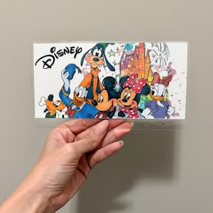 Könnte beinhalten: Ein klarer Kunststoffhalter mit einer farbenfrohen Illustration von Disney-Figuren, darunter Mickey Mouse, Minnie Mouse, Donald Duck und Goofy. Das Wort "Disney" ist in schwarzer Schrift über den Figuren geschrieben. Der Hintergrund zeigt ein Schloss und farbige Akzente.