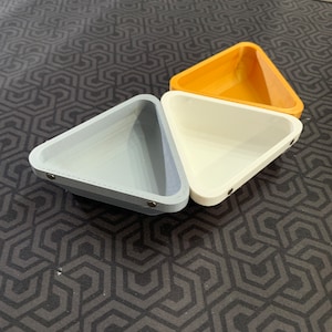 Puede incluir: Tres platos de forma triangular hechos de diferentes materiales. El plato de la izquierda es gris, el plato del medio es blanco y el plato de la derecha es naranja.