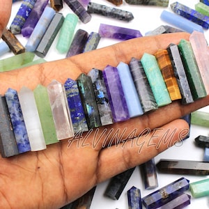 May include: A variety of gemstone wands in various colors, including blue, green, purple, and pink, are displayed in a hand. The wands are hexagonal and polished, with a pointed tip. The image includes the text "ALVINAGEMS".