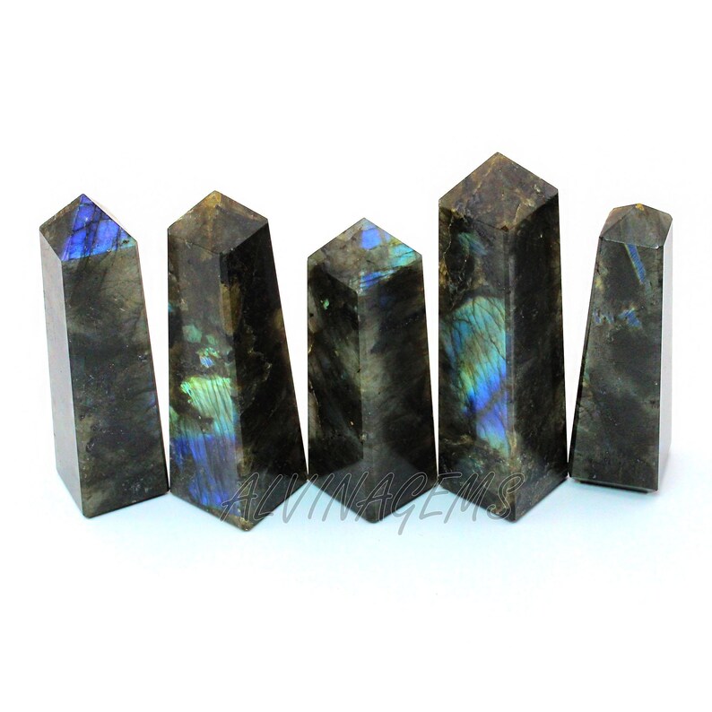 May include: Five polished labradorite crystal obelisks. The obelisks are dark gray with iridescent blue and green flashes. The obelisks are arranged in a row, each with a different height and shape. The text "ALVIN GEMS" is visible at the bottom.