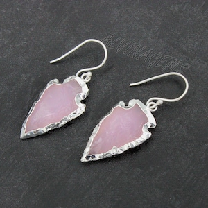 May include: A pair of earrings with a light pink arrowhead design. The earrings have a silver-colored metal border and ear hooks. The background is a dark gray color.