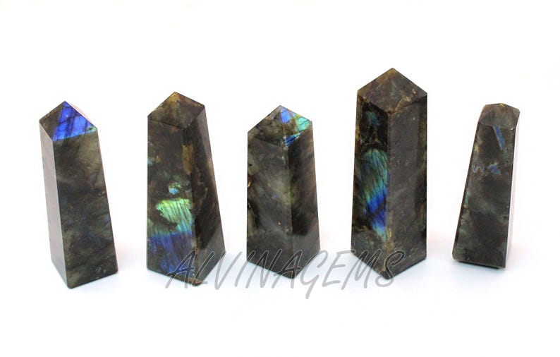 May include: Five polished labradorite crystal obelisks. The stones are dark gray with iridescent blue and green flashes. The obelisks are rectangular with a pointed top. The text "ALVINAGEMS" is visible on one of the obelisks.