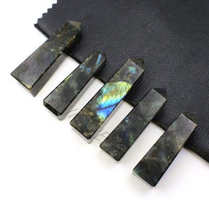 May include: Six rectangular labradorite crystal wands with angled tops. The wands are dark gray with iridescent blue, green, and yellow flashes. The wands are arranged on a black leather surface.