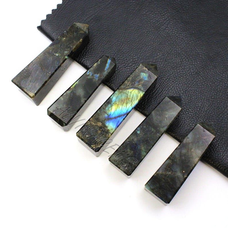 May include: Six rectangular labradorite crystal wands with angled tops. The wands are dark gray with iridescent blue, green, and yellow flashes. The wands are arranged on a black leather surface.