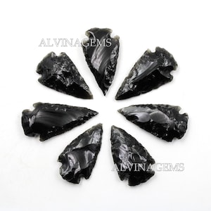 May include: Seven black obsidian arrowheads, each with a unique shape and texture, arranged in a circle on a white background. The arrowheads are approximately 5-7.5 cm in length. ALVINAGEMS is printed on the background.