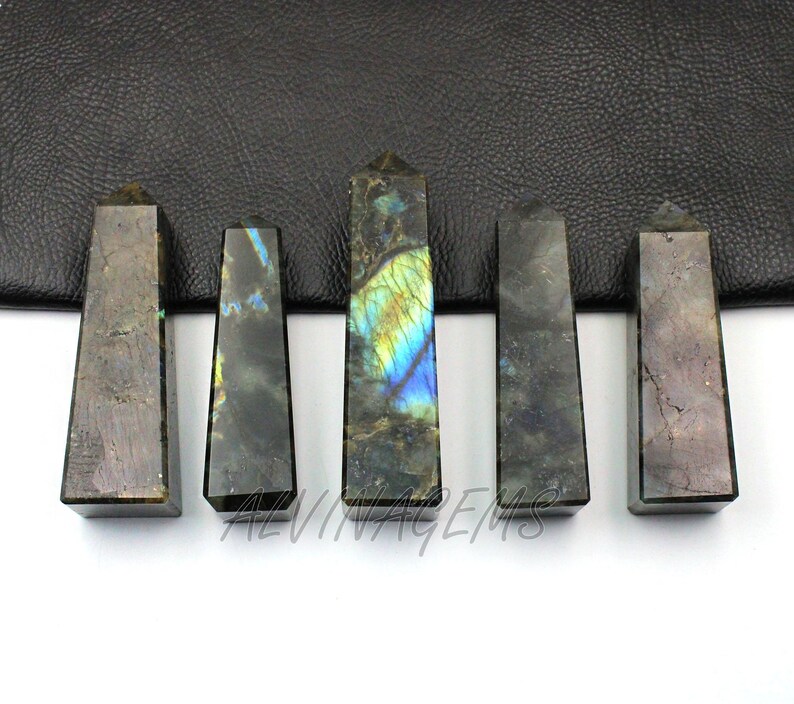 May include: Five polished labradorite obelisks. The obelisks are rectangular with a pointed top. The stones have a mix of gray, blue, and yellow colors. The text "ALVINTAGEEMS" is visible on the bottom.