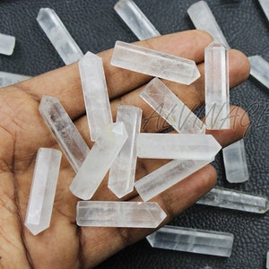 Może przedstawiać: Kolekcja przezroczystych kryształowych punktów kwarcu, znanych również jako różdżki, w różnych rozmiarach. Punkty są gładkie i wypolerowane, o przezroczystym wyglądzie. Kryształy są ułożone w dłoni, a niektóre z punktów wystają poza dłoń.