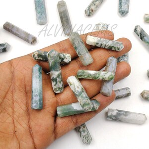 May include: A collection of polished, hexagonal-shaped gemstone crystals. The crystals are primarily white and gray with green inclusions, and are held in a hand. The crystals are of varying lengths.