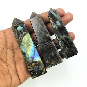 May include: Four rectangular labradorite crystal wands with pointed tips. The wands have a mix of dark gray, brown, and black colors with iridescent flashes of blue, green, and yellow. The wands are held in a hand.