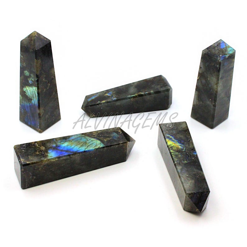 May include: Five polished labradorite crystal obelisks. The crystals are dark gray with iridescent blue, green, and yellow flashes. The obelisks vary in size and shape, with some standing upright and others lying horizontally.