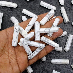 Puede incluir: Una colección de varitas de cristal de Howlita blanca con vetas grises. Las varitas son hexagonales y de varios tamaños. Los cristales están dispersos sobre una superficie oscura, algunos sostenidos en una mano. Estos cristales se utilizan a menudo para la curación y la meditación.