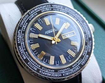 May include: A vintage wristwatch with a black leather band, displayed in a green box. The watch face is dark blue with gold-colored hands and markers. The bezel features city names in white text.