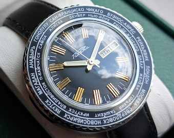 May include: A vintage wristwatch with a black leather band, featuring a dark face with gold-colored markers and hands. The watch has a silver bezel with city names and is presented in a green box.