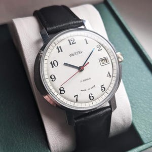 May include: A Wostok wristwatch with a white dial, black Arabic numerals, and a black leather strap. The watch features a date window at the three o'clock position, a red second hand, a blue minute hand, and the text 17 JEWELS and MADE IN USSR.