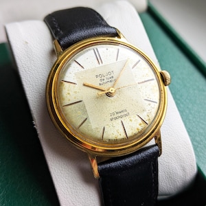 Puede incluir: Un reloj de pulsera vintage dorado con una correa de cuero negro. La esfera es de color crema claro con agujas y marcadores dorados. El texto "POLJOT de luxe automatic" y "29 jewels shockproof" es visible. El reloj está en una caja verde.