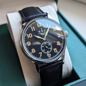 May include: A black leather wristwatch with a silver case and a black face. The watch has white numerals and a small second hand. The watch face also has the text "M.Ч.З. им. Кирова" in gold lettering.