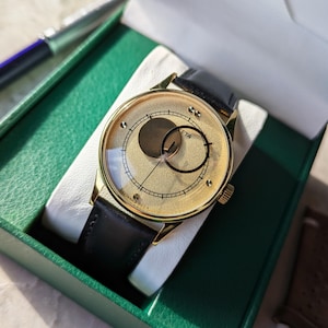 May include: A gold-tone wristwatch with a black leather band. The watch face has a unique design with two overlapping circles, one gold and one black, and the text "ETA" printed on the face.