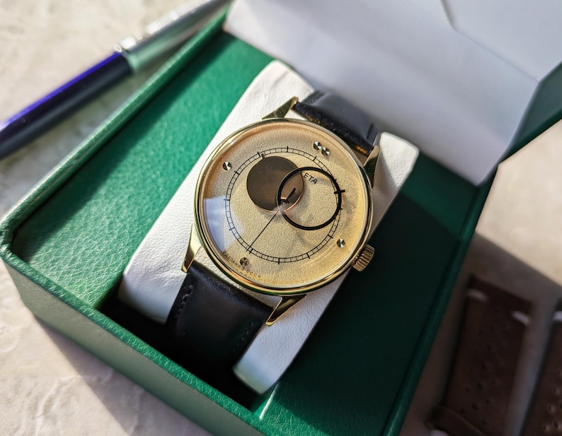 May include: A gold-tone wristwatch with a black leather band. The watch face has a unique design with two overlapping circles, one gold and one black, and the text "ETA" printed on the face.