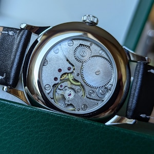 May include: A silver wristwatch with a black leather band. The back of the watch is open, revealing the intricate mechanical workings of the watch.