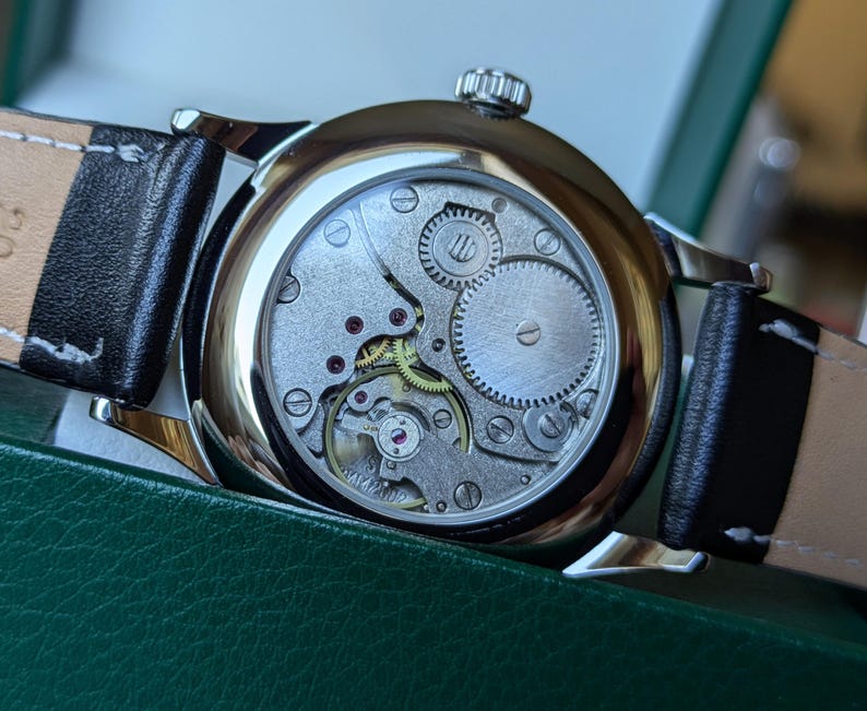 May include: A silver wristwatch with a black leather band. The back of the watch is open, revealing the intricate mechanical workings of the watch.