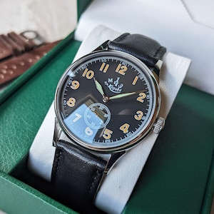 May include: A black and silver wristwatch with a black leather band. The watch face has white Arabic numerals and a small seconds dial. The watch face also has the text "м. Кирова" in orange.