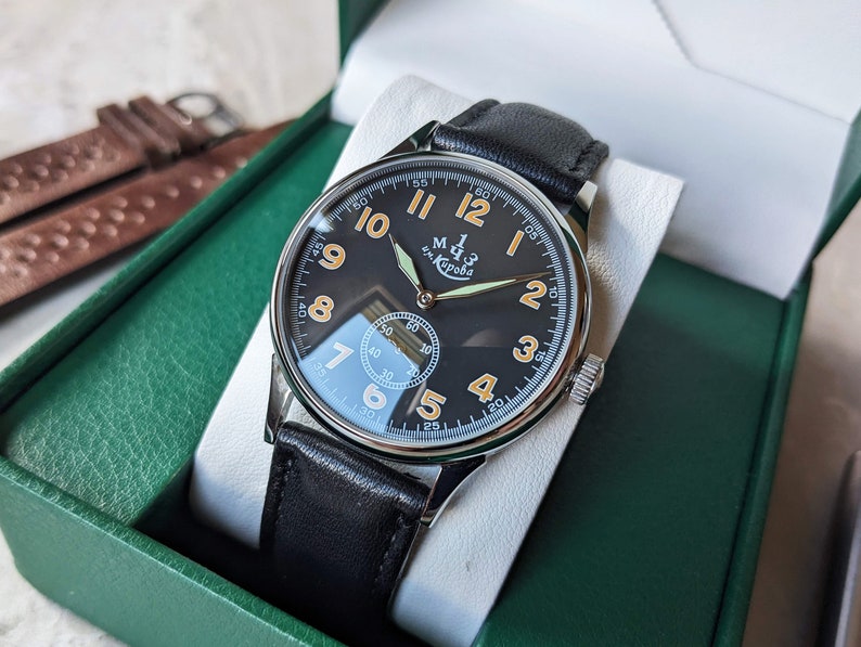 May include: A black and silver wristwatch with a black leather band. The watch face has white Arabic numerals and a small seconds dial. The watch face also has the text "м. Кирова" in orange.