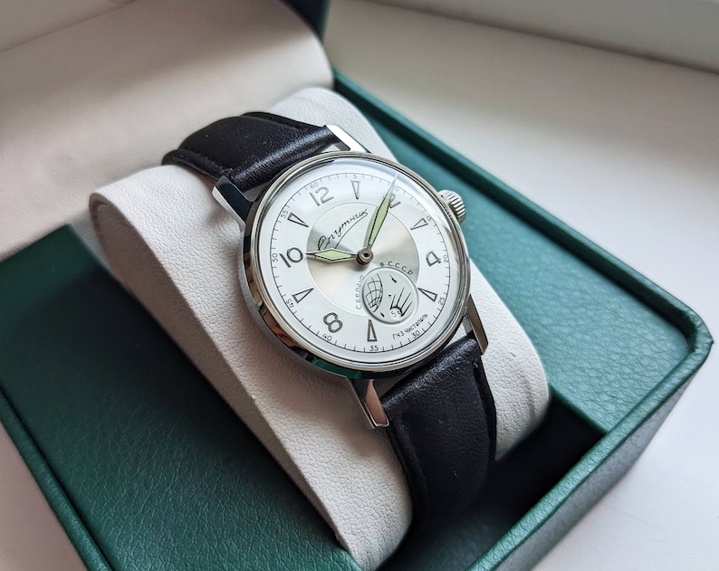 May include: A silver wristwatch with a white face and black leather band. The watch face has black Roman numeral hour markers and a small subdial at the 6 o'clock position. The watch face also features the text "СССР" and "Ракета" in Cyrillic script.