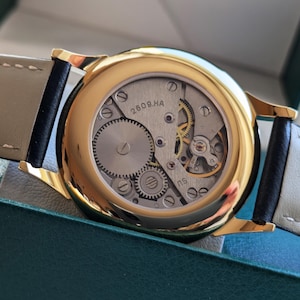 May include: A gold-tone wristwatch with a black leather band. The watch face is exposed, revealing the inner workings of the watch. The watch face has the text "2609.HA" printed on it.