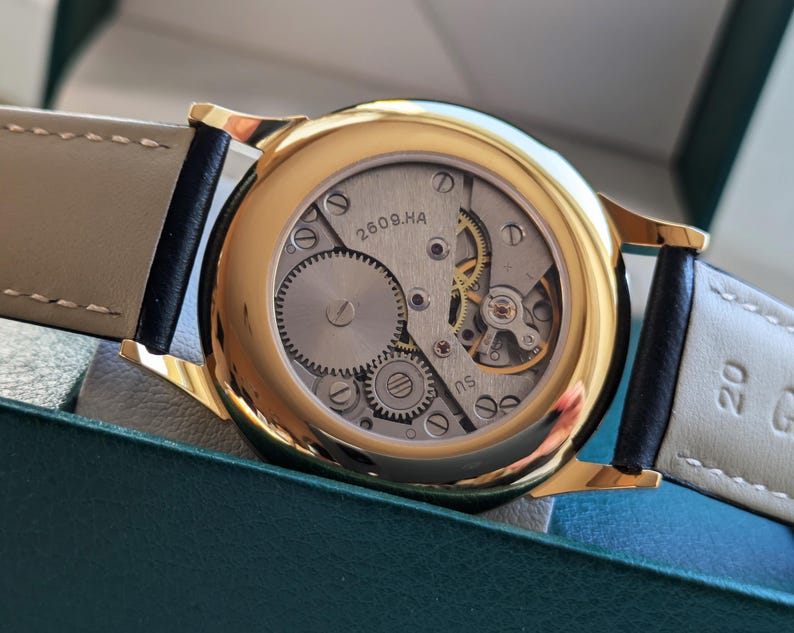 May include: A gold-tone wristwatch with a black leather band. The watch face is exposed, revealing the inner workings of the watch. The watch face has the text "2609.HA" printed on it.