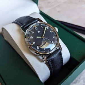 May include: A black leather wristwatch with a silver case and a black face. The watch has white numerals and a white second hand. The watch is in a green box.